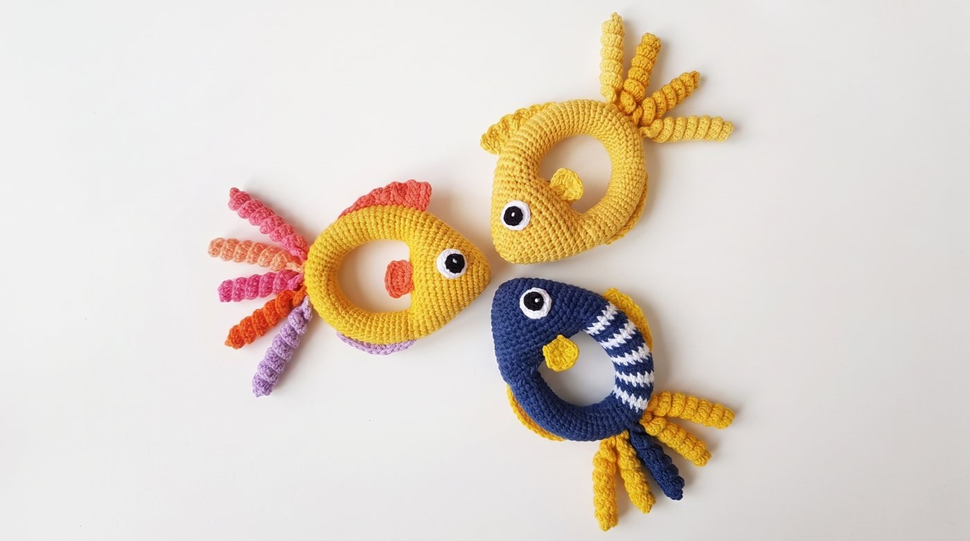 Drei gehäkelte Ring-Rasseln in Fischform in Gelb und Blau mit gewellten Flossen auf hellem Hintergrund