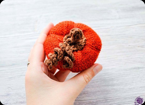Gestrickter orangeroter Kürbis von oben in der Hand mit braunen Lockenranken auf der Oberseite