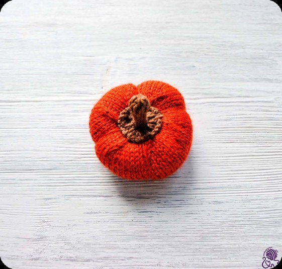 Gestrickter orangeroter Kürbis mit braunem Stiel von oben auf einem weißen Holztisch