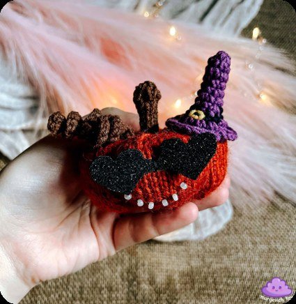 Gehäkelter und gestrickter Halloween-Kürbis in der Hand mit lila Hexenhut, schwarzer Herzbrille, Lockenranken und Perlenzähnen vor rosa Flausch