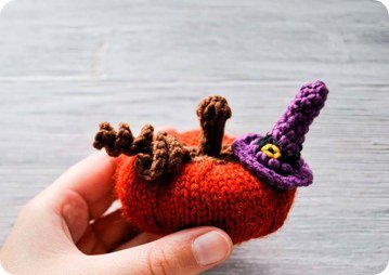 Halloween-Kürbis von der Seite in der Hand gehalten, mit braunem Stiel, Lockenranken und lila Hexenhut mit gelber Schnalle