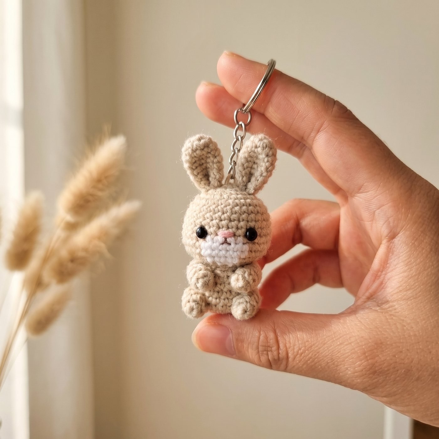 Beiger gehäkelter Hase als Schlüsselanhänger, an der Kette zwischen zwei Fingern gehalten, im Hintergrund Pampasgras